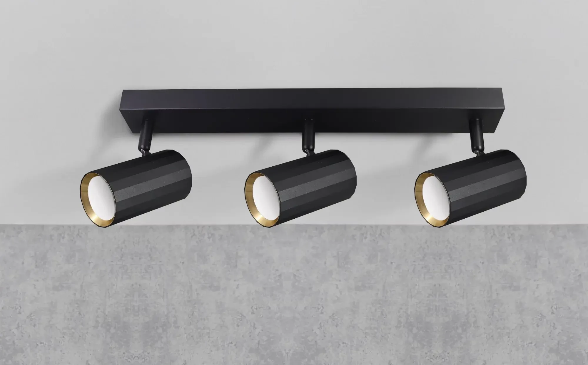 Lampa sufitowa Nicea z trzema reflektorami GU10 w kolorze czarnym i złotym. Ułożona równolegle do sufitu, z cylindrycznymi oprawami o teksturowanej powierzchni, z widocznymi białymi wnętrzami. Zainstalowana na prostokątnej, czarnej podstawie.