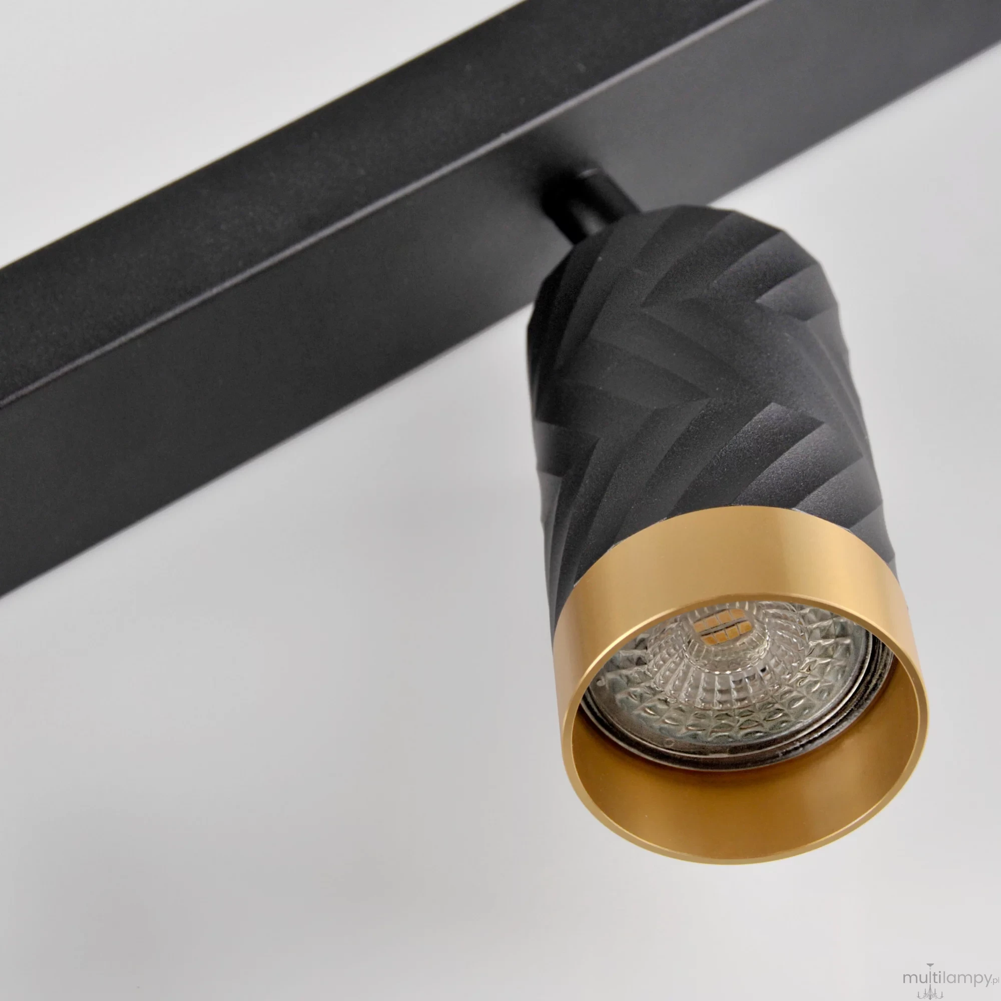 Lampa sufitowa Arras w kolorze czarnym i złotym, z dwoma reflektorami GU10. Widoczny detal w postaci teksturowanej obudowy reflektora oraz złotej obręczy. Ujęcie z bliska, ukazujące lampa montowaną na czarnej podstawie.