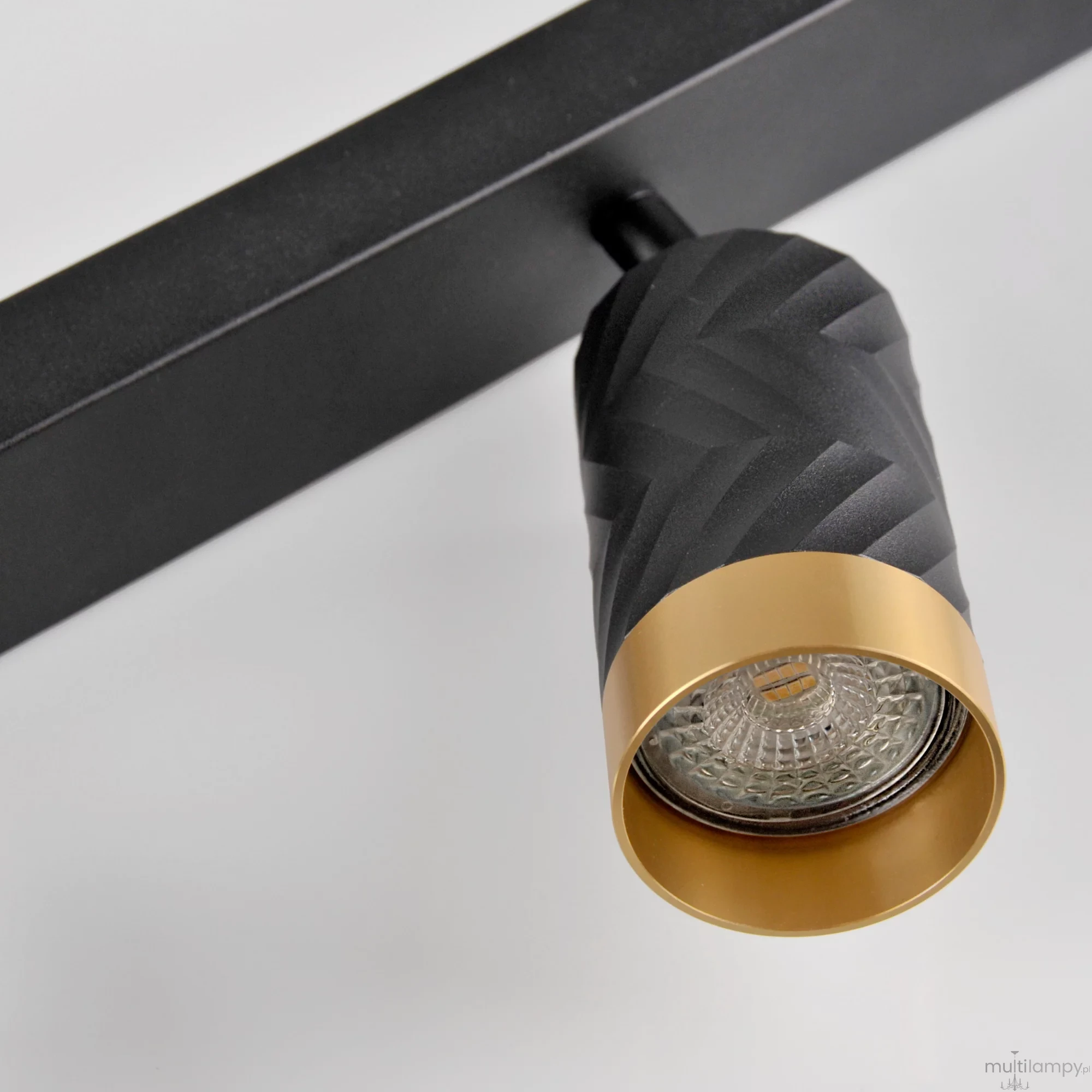 Lampa sufitowa Arras w kolorze czarnym i złotym z czterema reflektorami GU10. Kąt ujęcia zbliżenie na jeden reflektor o teksturowanej, czarnej obudowie z metalowym, złotym wykończeniem. Prosta, minimalistyczna konstrukcja na tle jasnego tła.