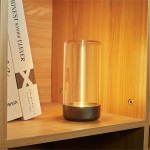 Esterno lampa stołowa z szarym, matowym podstawą i przezroczystym kloszem, o cylindrycznym kształcie. Zainstalowane LED 0,5W, IP44. Ujęcie z boku na półce z książkami, lampa włączona, emitująca ciepłe światło. Tło z drewnianych paneli.