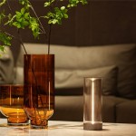 Stojąca lampa Esterno w kolorze szarym, z przezroczystym kloszem, z wbudowanym oświetleniem LED (1,2W), umieszczona na stole obok dekoracyjnych wazonów. IP44, odpowiednia do użytku na zewnątrz.