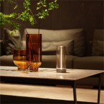 Lampa stołowa Esterno w kolorze szarym, z cylindrycznym korpusem, umieszczona na stole obok dwóch szklanych wazonów. Źródło światła LED o mocy 1,2W, z widocznym przyciskiem na podstawie. Tło stanowi miękka kanapa w odcieniu szarości.