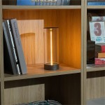 Lampa stołowa Esterno w kolorze szarym, z przezroczystym kloszem, emitująca ciepłe światło LED. Ustawiona na drewnianej półce wśród książek, z widocznym okrągłym podstawą. Wysokość lampy podkreśla nowoczesny design. IP44, 1xLED/1,2W.