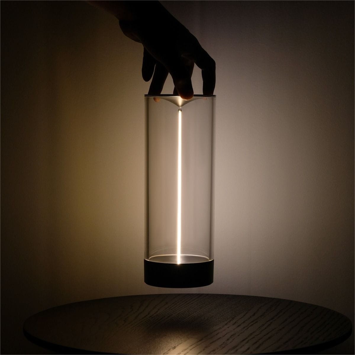 Lampa stołowa Esterno w szarym kolorze, z przezroczystym kloszem i LED-owym źródłem światła. Ujęcie z boku, ręka trzyma lampę nad okrągłym stołem. Widoczna smukła konstrukcja i emitowane światło tworzą minimalistyczny efekt.