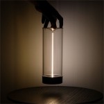 Lampa stołowa Esterno w szarym kolorze, z przezroczystym kloszem i LED-owym źródłem światła. Ujęcie z boku, ręka trzyma lampę nad okrągłym stołem. Widoczna smukła konstrukcja i emitowane światło tworzą minimalistyczny efekt.