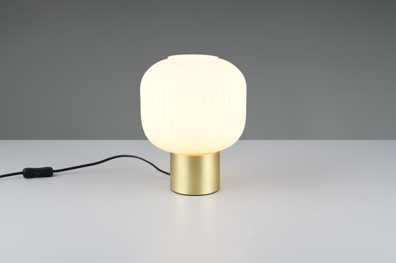 Lampa stołowa Diva 1xE27 w kolorze białym z mosiężną podstawą. Posiada zaokrągloną klosz z żłobioną fakturą, stojąca na okrągłej podstawie. Widoczny przewód z włącznikiem. Produkt na jednolitym tle.