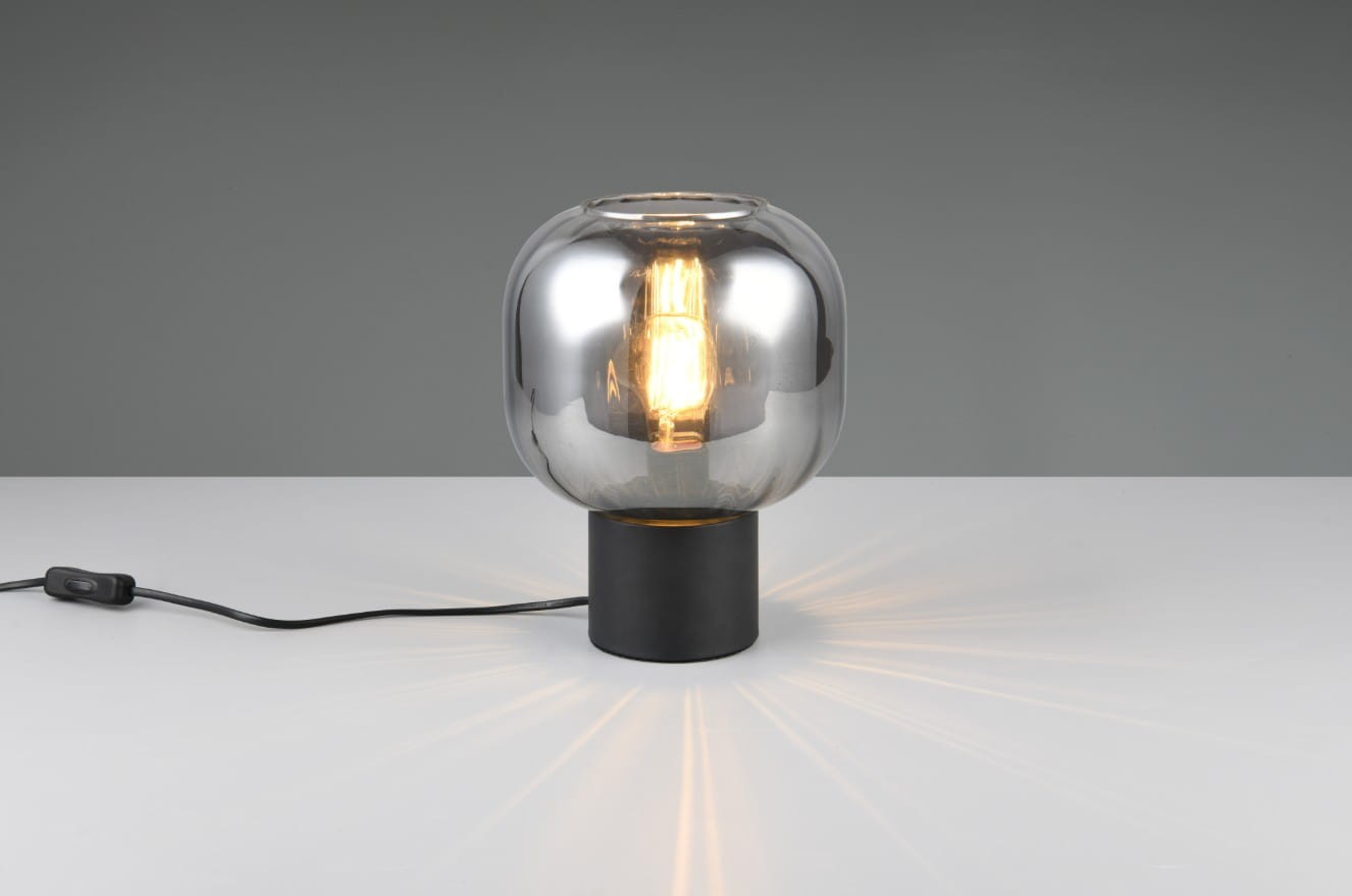 Lampa stołowa Diva z czarnym, okrągłym podstawą i chromowaną, szklaną kopułą. Widoczna żarówka E27 emitująca światło. Kabel z włącznikiem umiejscowiony z boku. Ujęcie frontalne, na jednolitym tle.