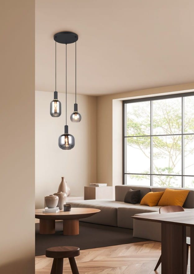 Lampa wisząca Diva w kolorze czarnym i chromowanym z trzema kloszami E27, zawieszona na długich przewodach. Widoczna w nowoczesnym wnętrzu z jasnymi ścianami, dużym oknem i meblami w odcieniach szarości oraz brązu.