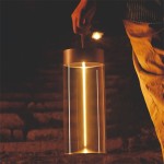 Lampa stołowa Esterno w kolorze szarym, z przezroczystym kloszem, trzymana w dłoni. Emituje ciepłe światło LED, o mocy 1,2W. Oświetlenie IP44, idealne do użytku na zewnątrz, w kontekście wieczornego otoczenia.