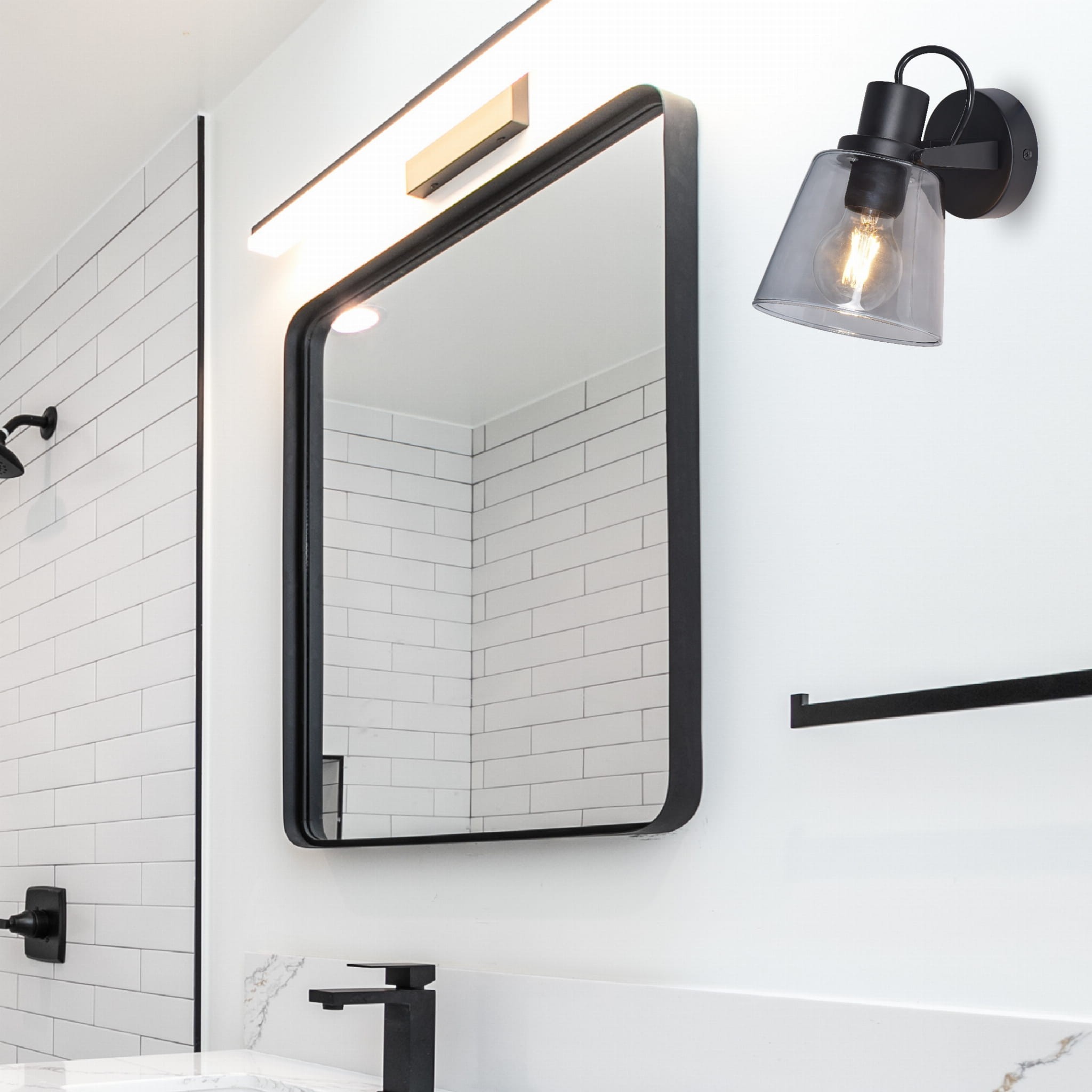 Kinkiet Badosa w kolorze czarnym z przezroczystym kloszem, montowany na ścianie. Zbliżenie na lampę przy lustrze w nowoczesnej łazience. Widoczne elementy: okrągła podstawa, metalowa konstrukcja oraz żarówka E27. Tło w jasnych odcieniach.