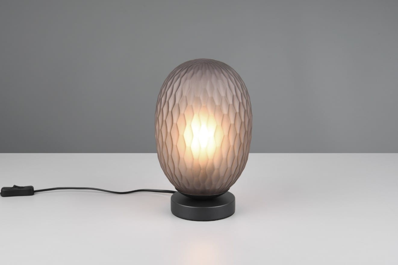 Lampa stołowa Facette w kolorze czarnym z przydymionym kloszem, o kształcie owalnym z fakturą w formie diamentu. Ustawiona na okrągłej podstawie, z widocznym kablem z włącznikiem. Jasne światło wydobywa się z wnętrza klosza.