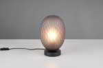 Lampa stołowa Facette w kolorze czarnym z przydymionym kloszem, o kształcie owalnym z fakturą w formie diamentu. Ustawiona na okrągłej podstawie, z widocznym kablem z włącznikiem. Jasne światło wydobywa się z wnętrza klosza.