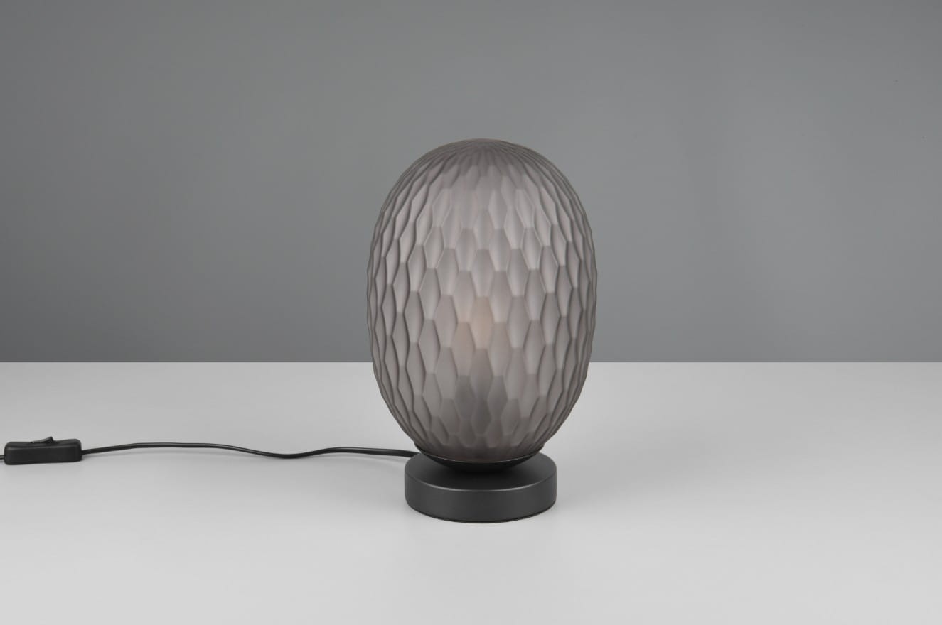 Lampa stołowa Facette z czarnym, okrągłym stelażem i przydymionym kloszem o kształcie owalnym z fakturą w formie siatki. Umieszczona na białym stole, z kablem zasilającym widocznym z boku.