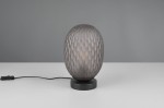 Lampa stołowa Facette z czarnym, okrągłym stelażem i przydymionym kloszem o kształcie owalnym z fakturą w formie siatki. Umieszczona na białym stole, z kablem zasilającym widocznym z boku.