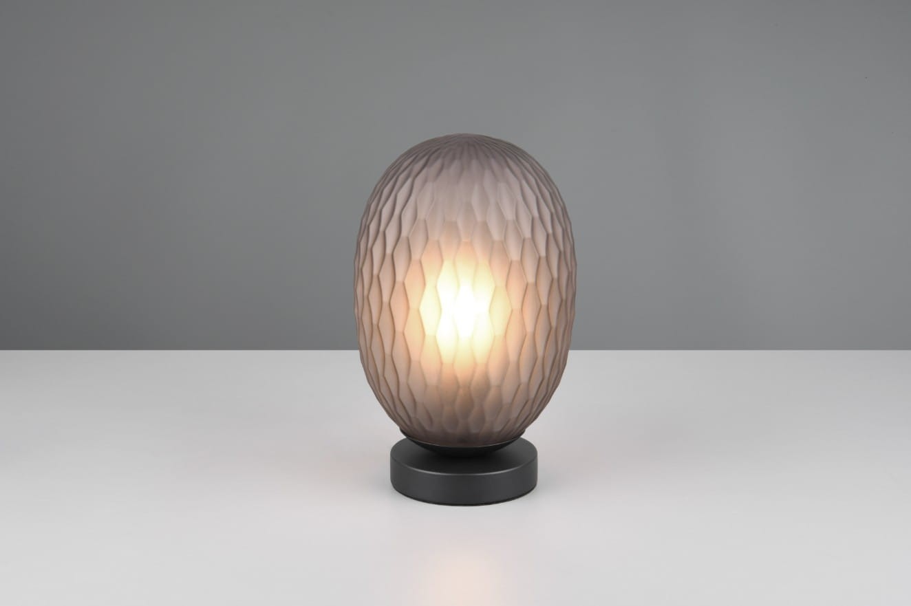 Lampa stołowa Facette z kloszem w kształcie elipsoidy, wykonanym z przydymionego szkła z fakturą w formie diamentowych wzorów, umieszczona na czarnej, okrągłej podstawie. Oświetlenie z żarówką E27.