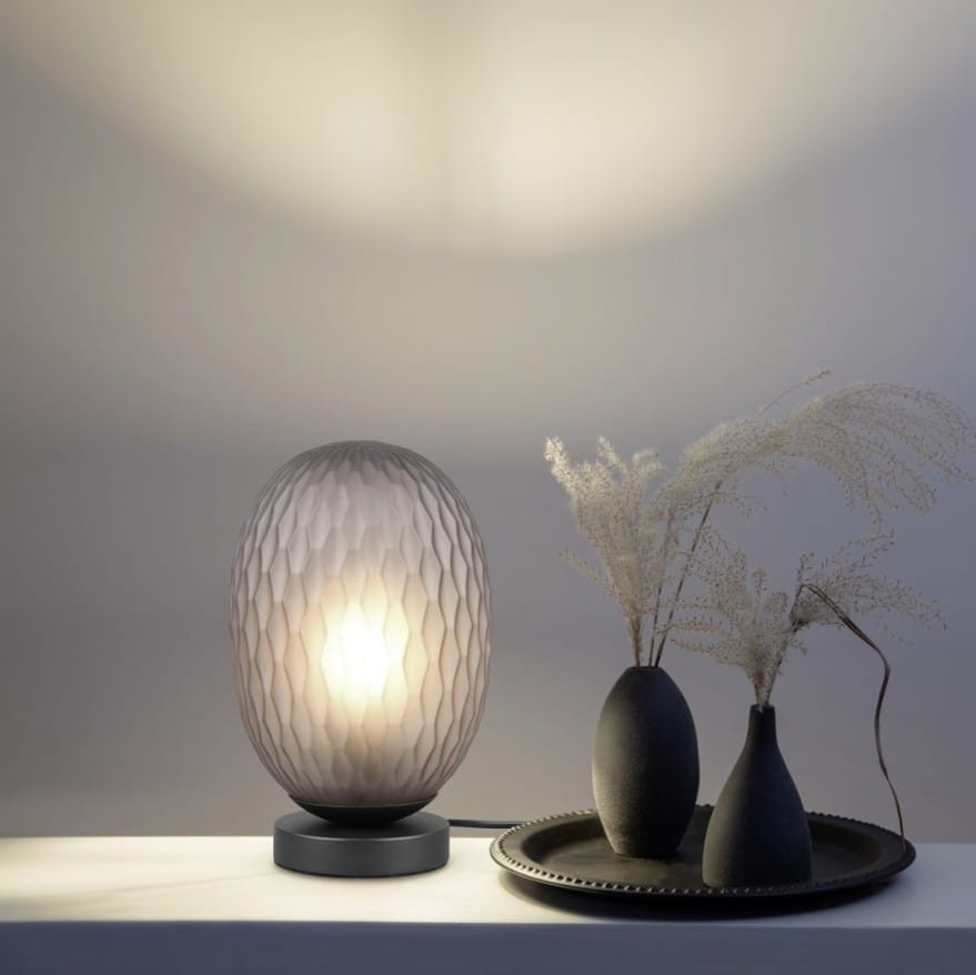 Lampa stołowa Facette w kolorze czarnym z przydymionym, owalnym kloszem o fakturze w drobne żłobienia. Stoi na okrągłej podstawie, obok dekoracyjne wazy i talerz. Tło jasne, minimalistyczne.