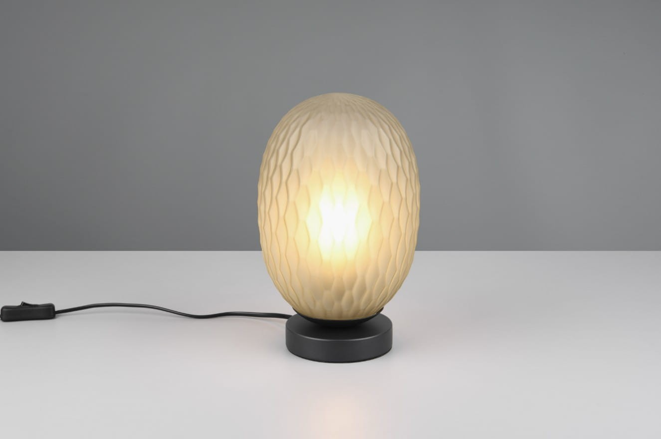 Lampa stołowa Facette z kloszem w kształcie elipsoidy w kolorze białym z teksturą w formie rombów, osadzona na czarnej podstawie. Widoczny przewód z włącznikiem. Oświetlenie włączone, emitujące ciepłe światło.