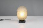 Lampa stołowa Facette z kloszem w kształcie elipsoidy w kolorze białym z teksturą w formie rombów, osadzona na czarnej podstawie. Widoczny przewód z włącznikiem. Oświetlenie włączone, emitujące ciepłe światło.