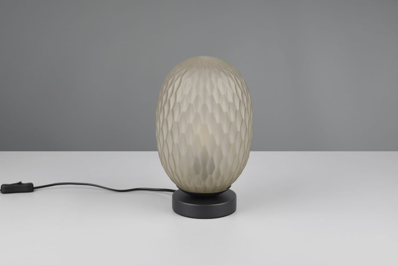 Lampa stołowa Facette z kloszem w kształcie owalnym, pokrytym strukturą w formie rombów, w kolorze mlecznobiałym, osadzona na czarnej, okrągłej podstawie. Widoczny kabel z włącznikiem. Ujęcie frontalne.