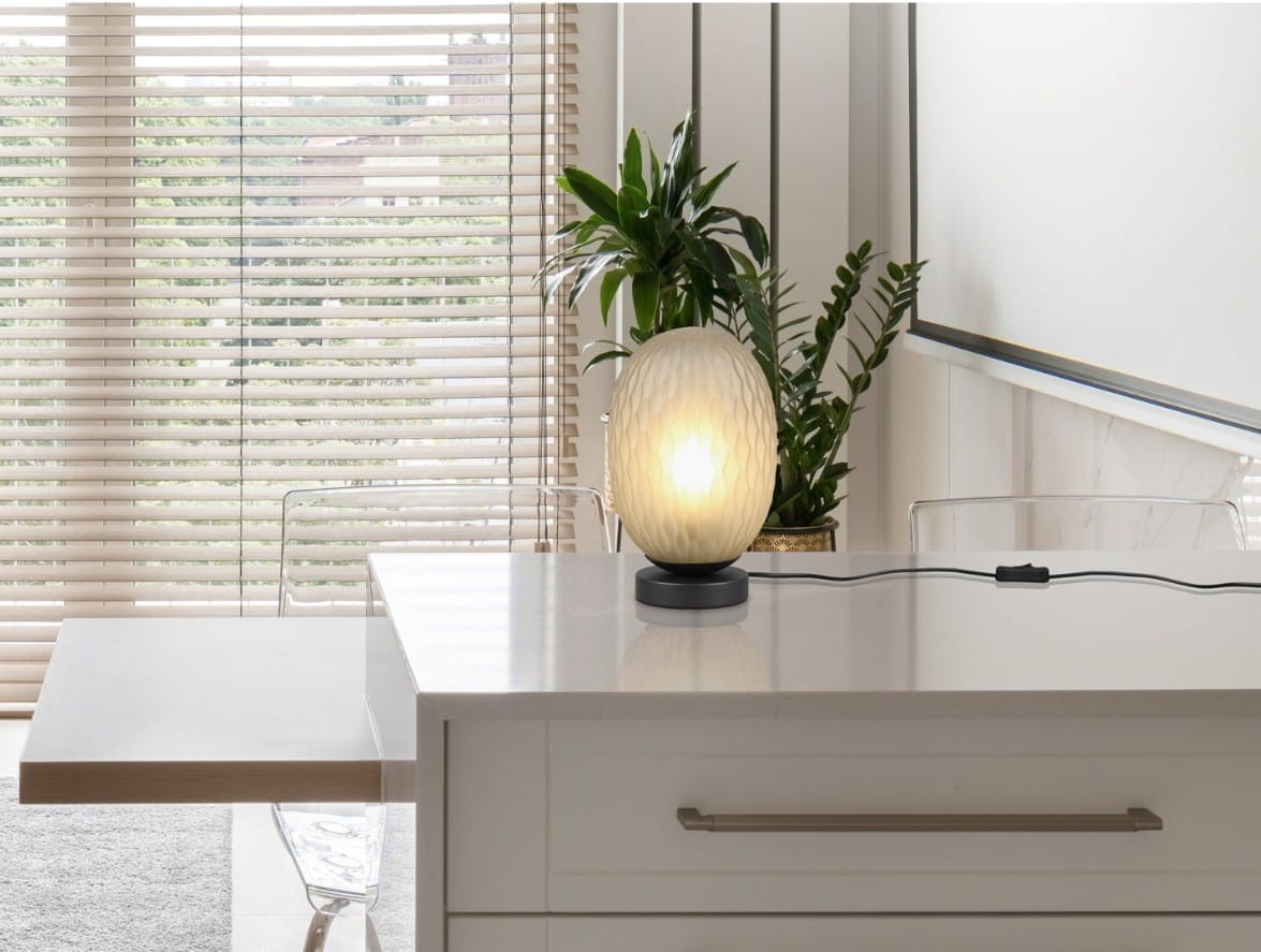 Lampa stołowa Facette z oprawą w czerni i zieleni, z żarówką E27. Owalna, teksturowana klosz w jasnym kolorze, umieszczona na czarnej podstawie. Widoczny kabel z włącznikiem, lampa ustawiona na białym blacie w jasnym wnętrzu z roślinami w tle.