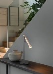 Lampa stołowa Fiato w kolorze czarnym i złotym, z regulowanym ramieniem. Znajduje się na ciemnym blacie obok ceramicznego dzbanka. Tło stanowi szara ściana oraz roślinność, co tworzy minimalistyczną aranżację.
