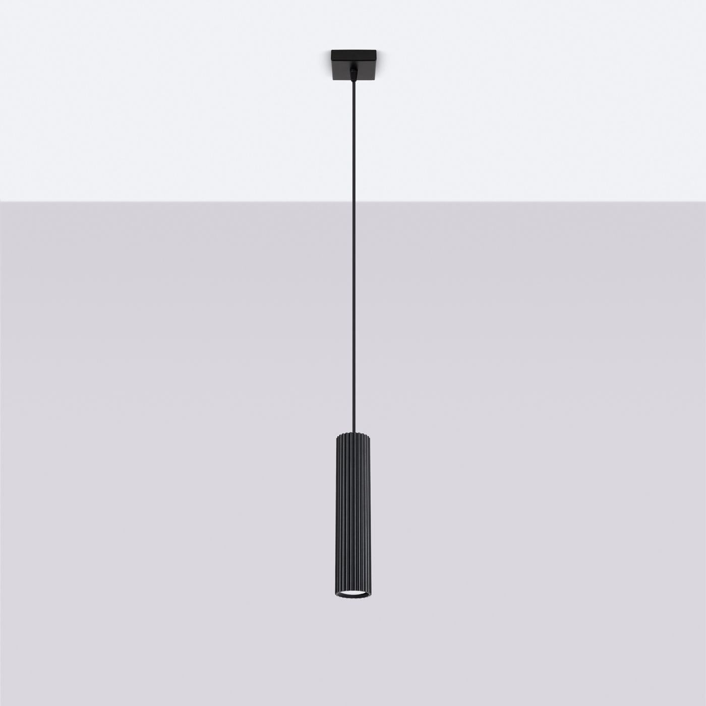 Lampa wisząca Karbon 1 o czarnej, cylindrycznej obudowie z rowkowaniem. Ujęcie frontalne, zawieszona na cienkim kablu. Montaż sufitowy, z oprawką na żarówkę GU10. Tło w jasnym odcieniu szarości, podkreślające produkt.