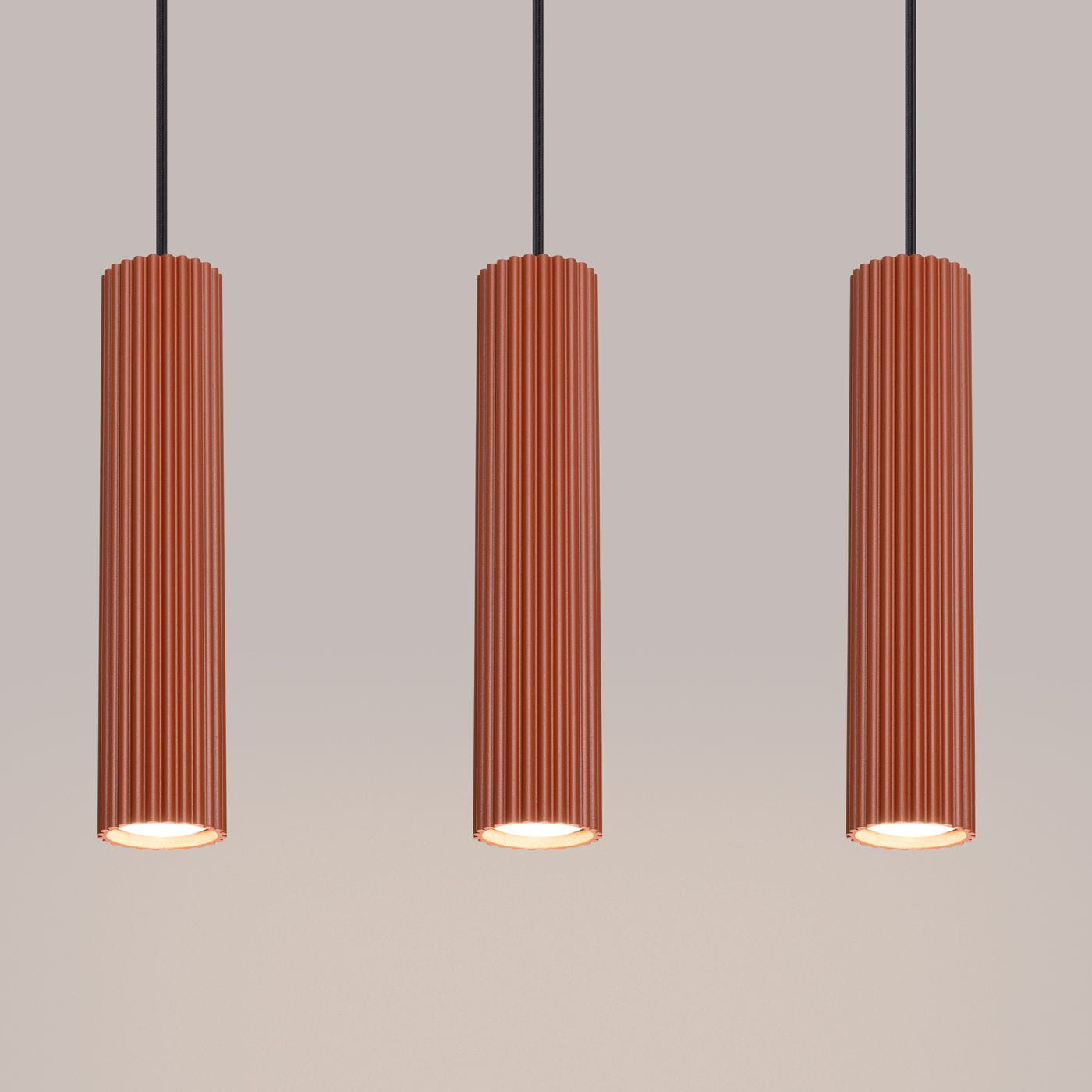 Lampa wisząca Karbon 3, w kolorze ochra czerwona, składająca się z trzech cylindrycznych kloszy z pionowymi żłobieniami. Klosze zawieszone na czarnych przewodach, emitujące światło z trzech opraw GU10. Tło jasne, jednolite.