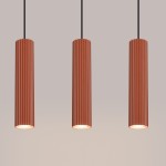 Lampa wisząca Karbon 3, w kolorze ochra czerwona, składająca się z trzech cylindrycznych kloszy z pionowymi żłobieniami. Klosze zawieszone na czarnych przewodach, emitujące światło z trzech opraw GU10. Tło jasne, jednolite.