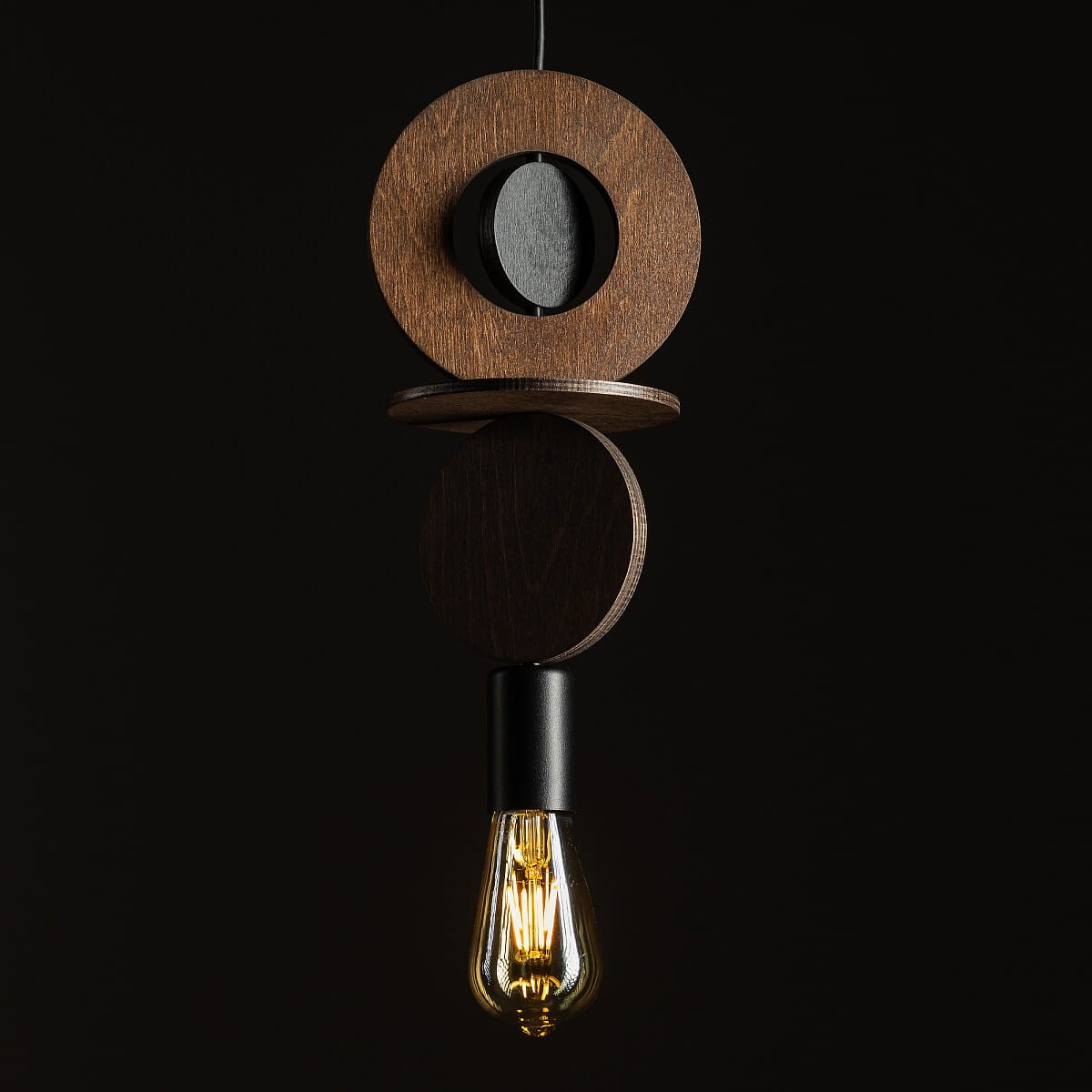 Lampa wisząca Drops Wood C z jednym gniazdem E27. Składa się z okrągłych elementów z ciemnego drewna oraz czarnego detalu. W dolnej części umieszczona żarówka o vintage''owym kształcie. Produkt zawieszony na cienkim przewodzie, na jednolitym ciemnym tle.