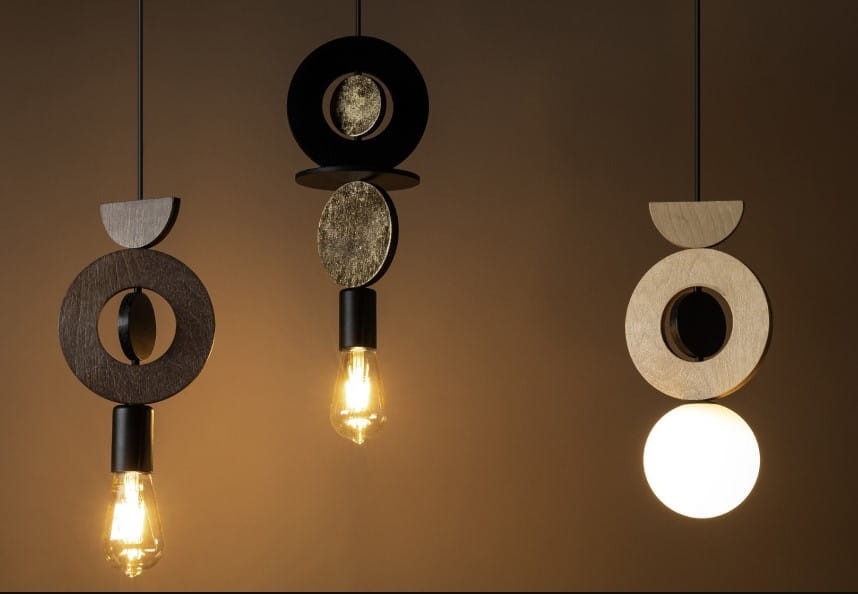 Lampa wisząca Drops Wood F z trzech elementów: czarna okrągła część z metalowym wykończeniem, złoty detal oraz jasne drewniane akcenty. Źródło światła E27, widoczny jeden żarówka, umieszczona w dolnej części. Tło w odcieniach brązu.