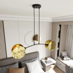 Lampa wisząca Motif w kolorze złotym i czarnym, z dwoma okrągłymi kloszami o fakturze przypominającej marmur. Ułożona w architektoniczny kształt, zwisająca z sufitu na dwóch kablach. Tło przedstawia nowoczesne wnętrze sypialni z łóżkiem i dekoracjami.