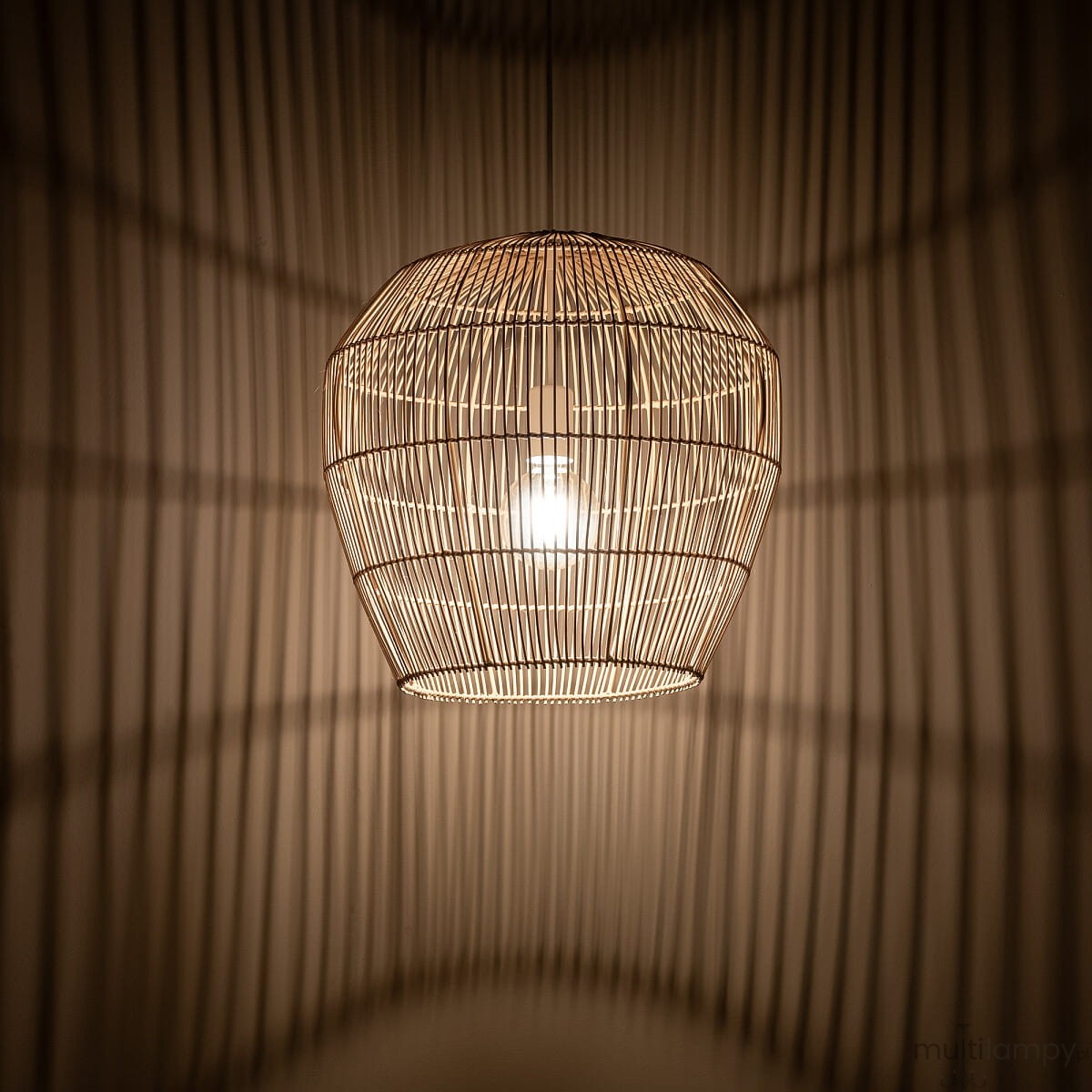 Lampa wisząca Haiti S, w białym i naturalnym kolorze, wykonana z cienkich prętów, o owalnym kształcie. Zawieszona na suficie, emituje ciepłe światło, rzucając delikatny cień na ścianę. Wnętrze lampy widoczne, z żarówką E27 w środku.