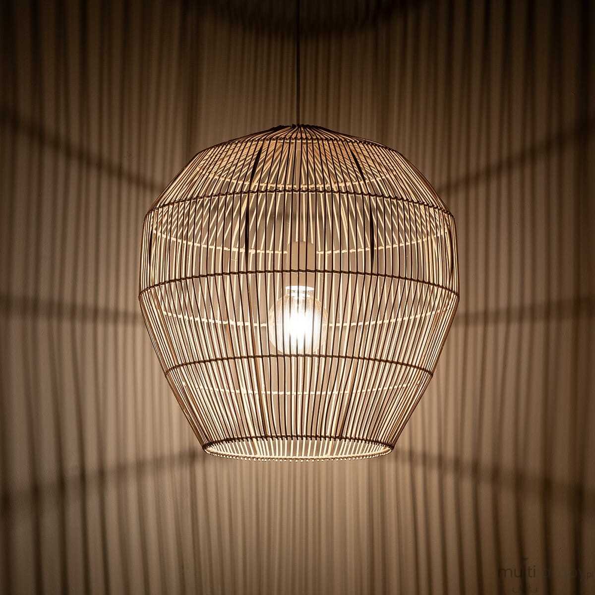 Lampa wisząca Haiti w białym i naturalnym kolorze, o kształcie heksagonalnym, wykonana z cienkich prętów. Widoczna żarówka E27 wewnątrz, tworząca delikatne cienie na ścianie. Zawieszona na cienkim przewodzie, w górnej części pomieszczenia.