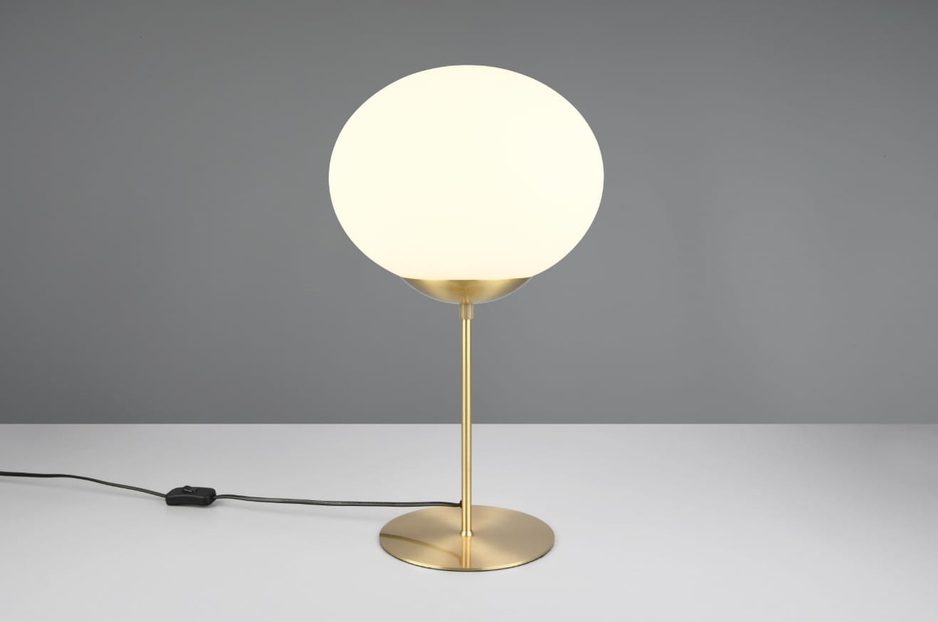 Lampa stołowa Fomento z mosiężną podstawą i białym, okrągłym kloszem. Ujęcie frontalne, lampa stoi na białym blacie. Widoczny czarny kabel z przełącznikiem. Prosta forma, eleganckie wykończenie.