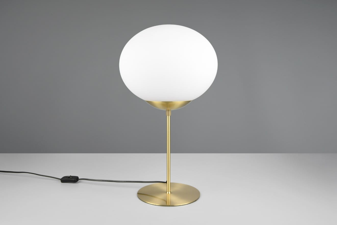 Lampa stołowa Fomento 1xE27 w kolorze mosiądzu z białym, okrągłym kloszem. Produkt stoi na okrągłej podstawie, z widocznym kablem z włącznikiem. Tło w odcieniu szaro-beżowym, podkreślające elegancki design lampy.