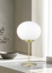 Lampa stołowa Fomento z mosiężną podstawą i białym, okrągłym kloszem. Ujęcie z przodu, stojąca na jasnym blacie. Widoczne przewody. Prosta forma, elegancki design, idealna do nowoczesnych wnętrz.