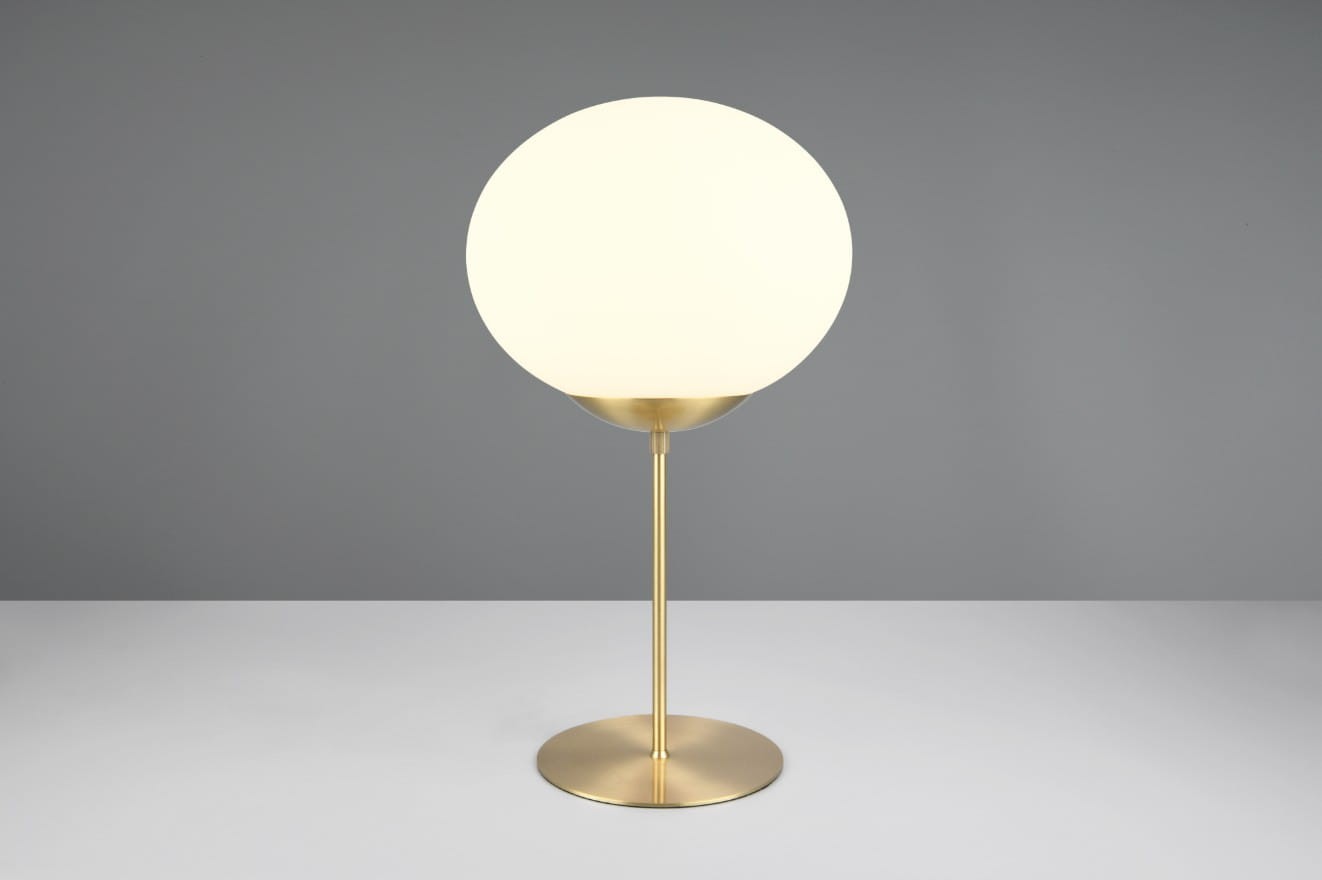 Lampa stołowa Fomento z mosiężną podstawą i białym, okrągłym kloszem. Ujęcie frontalne, lampa stoi na płaskiej powierzchni. Klosz emituje miękkie światło, a elegancka konstrukcja podkreśla nowoczesny design. Tło w odcieniu szarym.