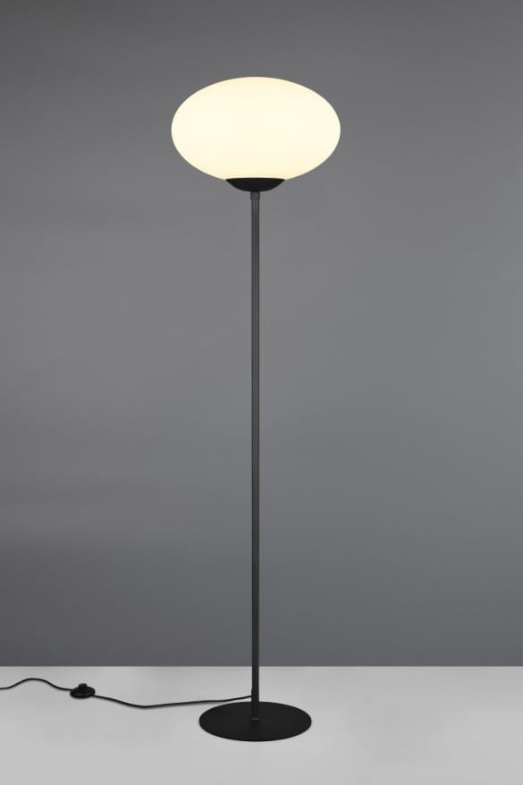 Lampa podłogowa Fomento w kolorze czarnym i białym, z okrągłym, białym kloszem. Stojąca, z metalową podstawą i smukłym stelażem. Widoczny przewód z wtyczką na podłodze. Ujęcie frontalne na jednolitym tle.
