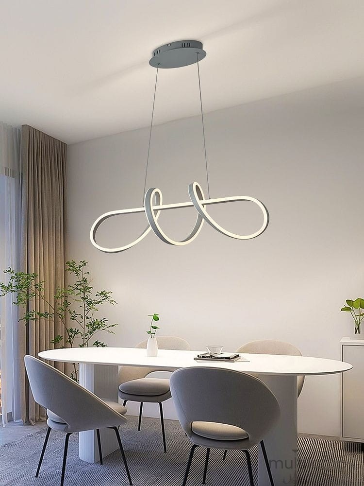 Lampa wisząca Trevance 1xLED/56W w kolorze szarym, o nowoczesnym, organicznym kształcie, zawieszona na cienkich linkach. Umieszczona nad okrągłym stołem w jasnym wnętrzu, z roślinnością w tle.