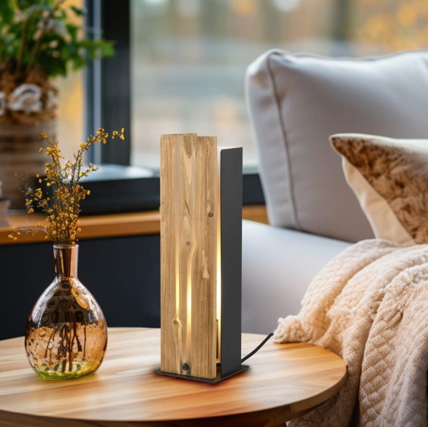 Lampa stołowa Karuni z jedną oprawą E27, w kolorze czarnym i jasnego drewna. Prosta forma z drewnianym elementem i metalową podstawą. Zbliżenie na lampę umieszczoną na drewnianym stole obok dekoracyjnej butelki i kwiatów.