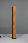 Lampa podłogowa Karuni z czarnym korpusem i jasnym drewnem, wąska, o prostym, pionowym kształcie. Widoczne ciepłe światło przez szczeliny w drewnie. Stoi na płaskiej podstawie, z przewodem elektrycznym po lewej stronie. Tło szare.