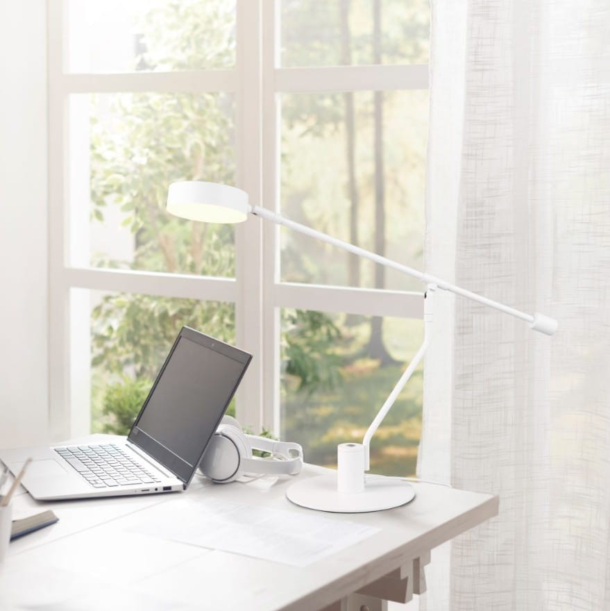 Biała lampa biurkowa Manduro z jednym źródłem LED 6W, z ruchomym ramieniem i okrągłą podstawą. Ujęcie z boku na biurku obok laptopa i słuchawek, w jasnym wnętrzu z dużymi oknami w tle.
