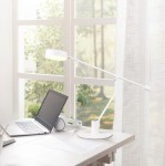 Biała lampa biurkowa Manduro z jednym źródłem LED 6W, z ruchomym ramieniem i okrągłą podstawą. Ujęcie z boku na biurku obok laptopa i słuchawek, w jasnym wnętrzu z dużymi oknami w tle.