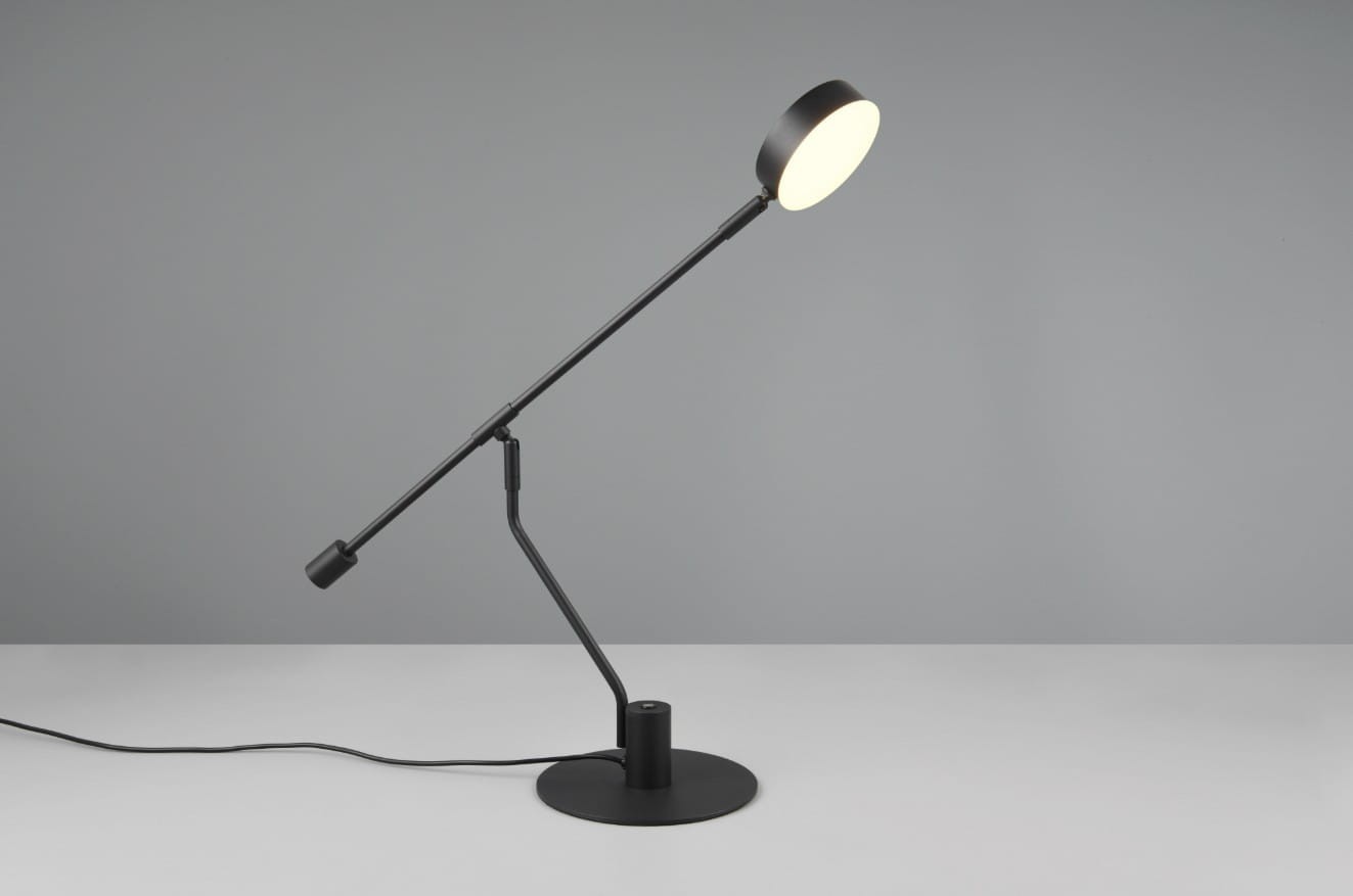 Czarna lampa biurkowa Manduro z jednym źródłem LED 6W, ustawiona w pozycji stojącej. Posiada regulowany ramie oraz okrągłą podstawę, z okrągłą głowicą emitującą światło. Tło w odcieniach szarości, prosty design.