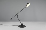 Czarna lampa biurkowa Manduro z jednym źródłem LED 6W, ustawiona w pozycji stojącej. Posiada regulowany ramie oraz okrągłą podstawę, z okrągłą głowicą emitującą światło. Tło w odcieniach szarości, prosty design.