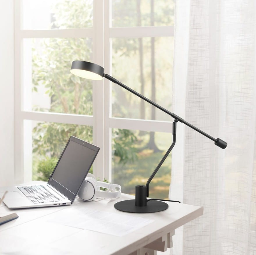 Czarna lampa biurkowa Manduro z regulowanym ramieniem, z jedną diodą LED 6W. Ustawiona na biurku obok laptopa, na płaskiej podstawie. Tło stanowi jasne wnętrze z oknem, wpuszczającym naturalne światło.