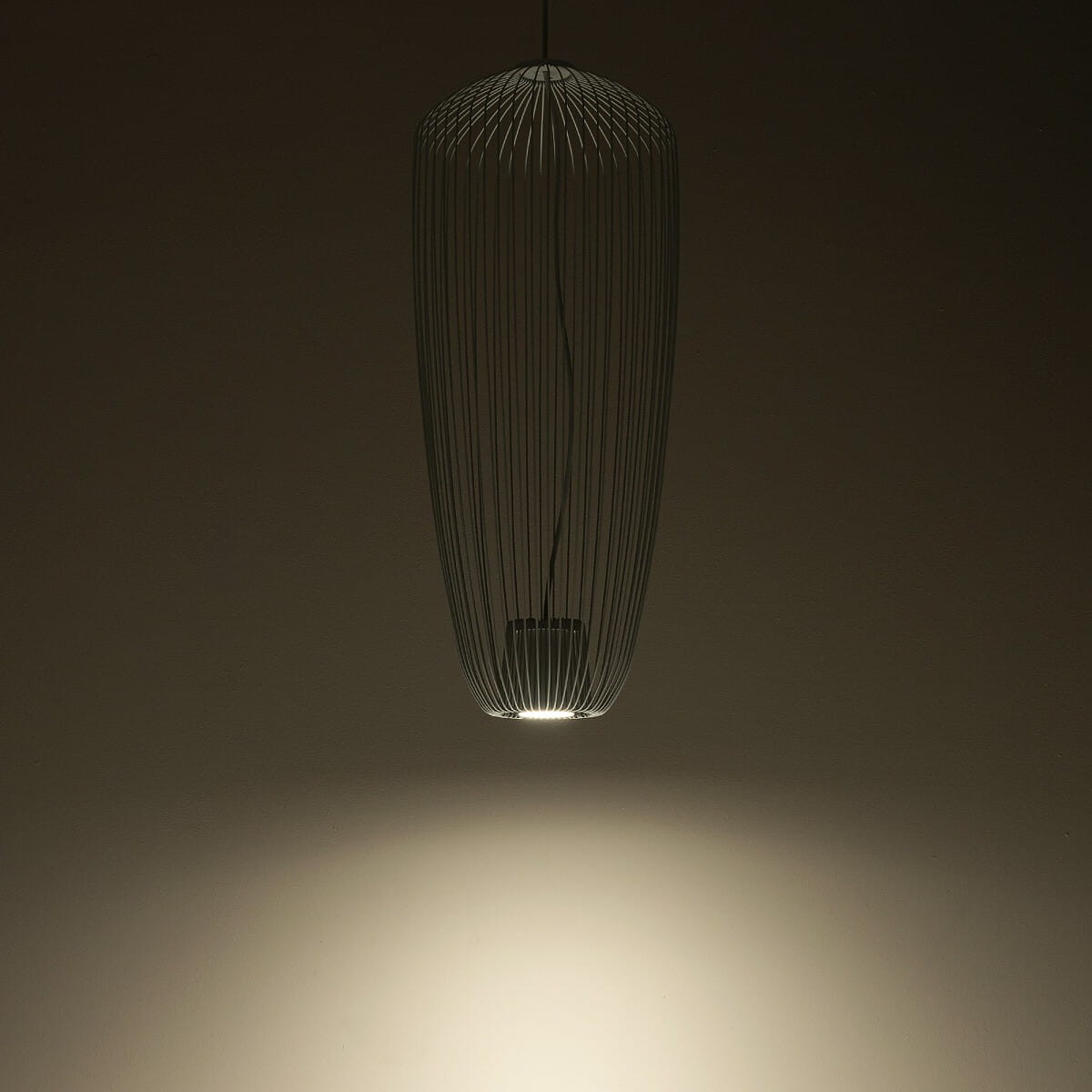 Lampa wisząca Pumpkin S w kolorze jasnoszarym o kształcie stożkowatym z cienkimi, metalowymi prętami. Zawieszona na cienkim przewodzie, z widoczną żarówką GU10 emitującą światło w dół, na tle ciemnej ściany.