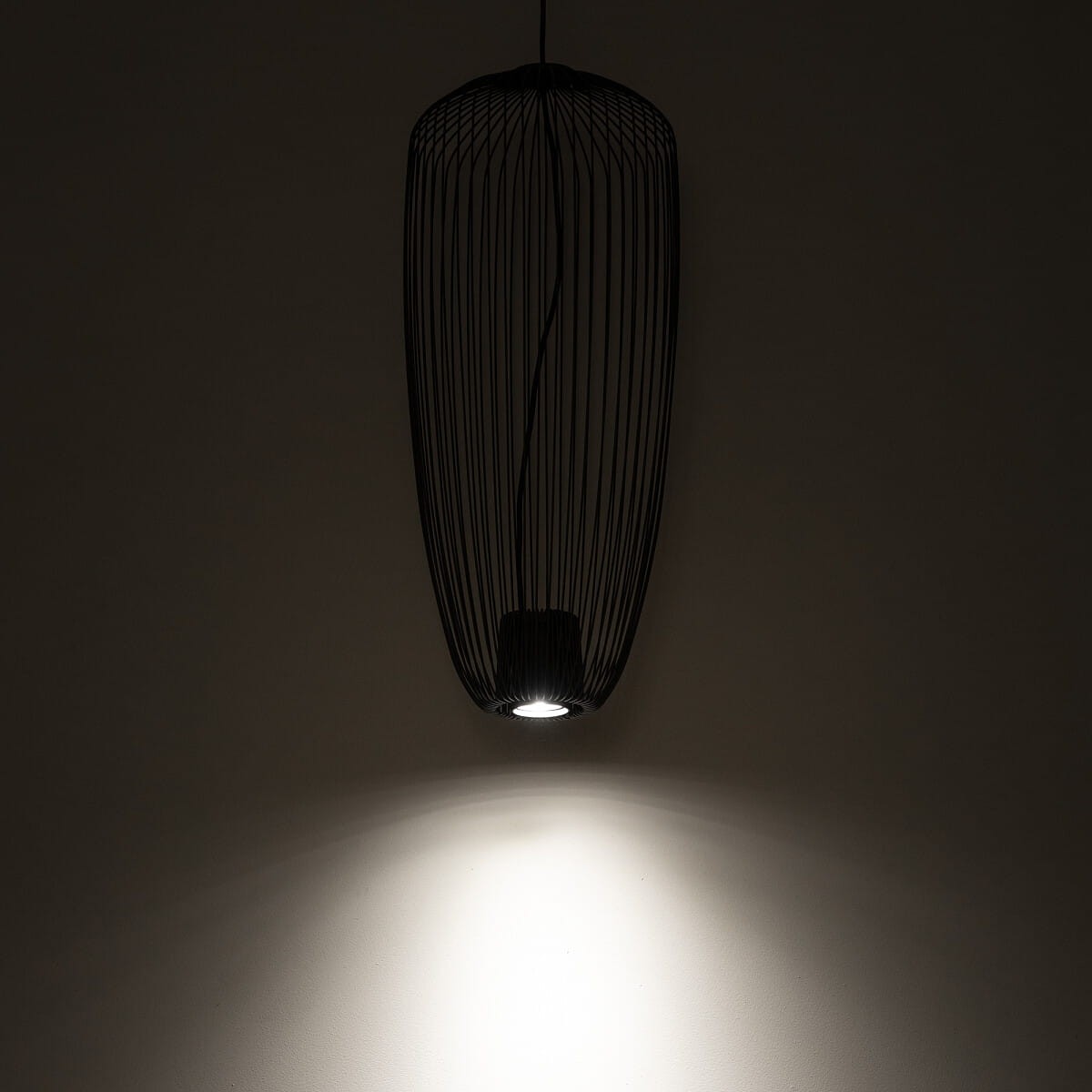 Lampa wisząca Pumpkin S w kolorze szarym, z metalową siatką w kształcie owalnym, zawieszona na cienkim przewodzie. Źródło światła GU10 w dolnej części, emitujące jasne światło na ścianę. Tło ciemne, podkreślające design lampy.