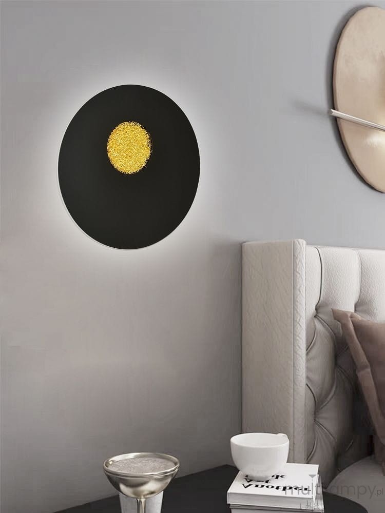 Kinkiet Savelo 2xLED/24W w kolorze czarnym, okrągły kształt z centralnym, złotym detalem. Zawieszony na ścianie, w tle jasna ściana i nowoczesne meble. Widoczna elegancka srebrna miska na stoliku obok.