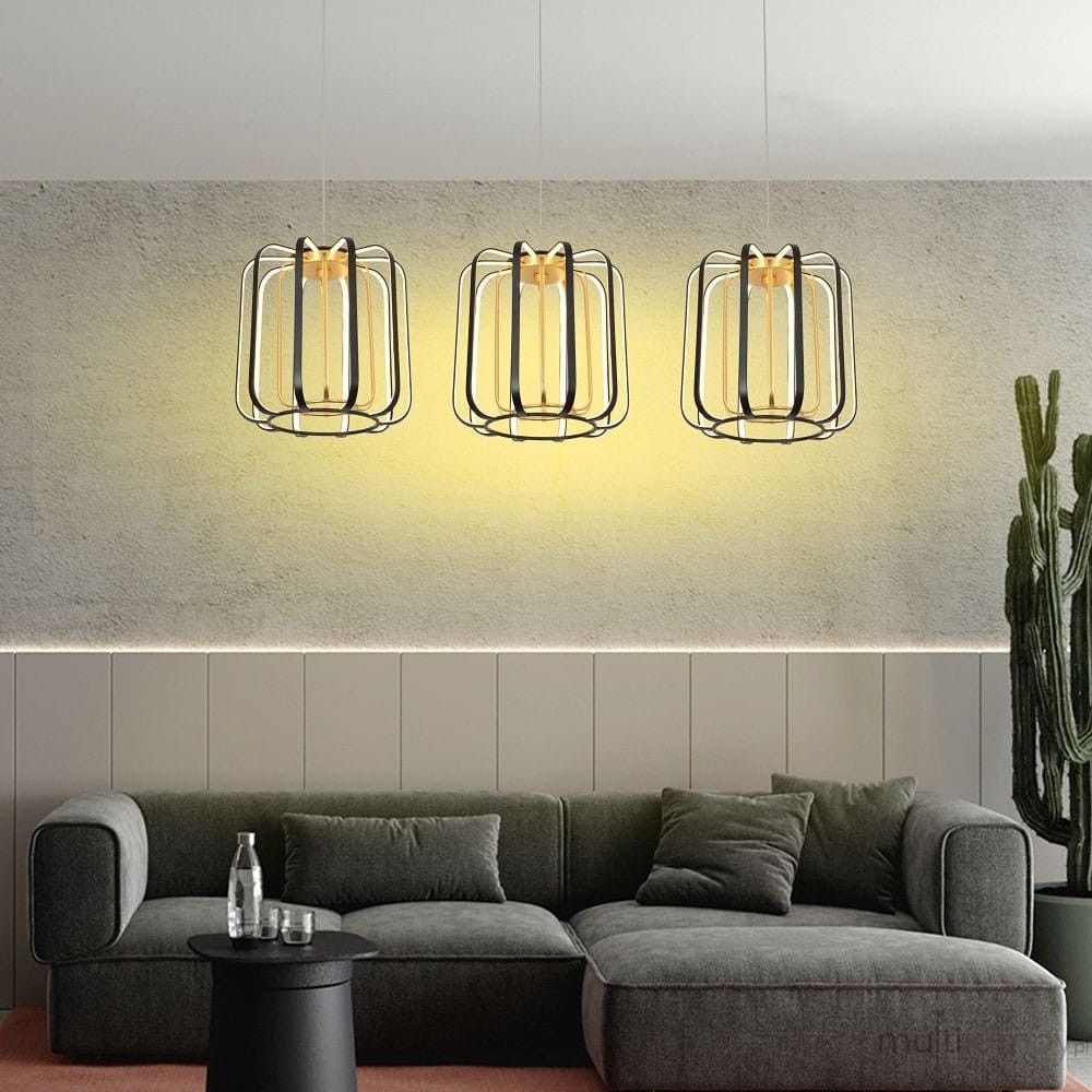 Lampa wisząca Xara z trzema źródłami LED, w kolorze czarnym z elementami złotymi. Oświetlenie ma nowoczesny design, zawieszona w rzędzie nad szaro-zieloną sofą, na tle jasnej, surowej ściany.